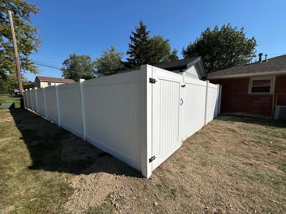White vinyl fence enclosing yard with gate, surrounded by green grass and trees.