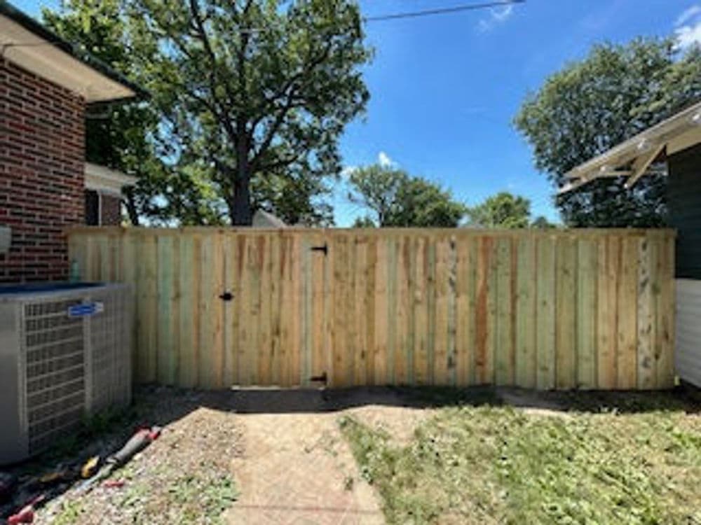 Wooden privacy fence with a gate, surrounded by grass and trees, under a clear blue sky.