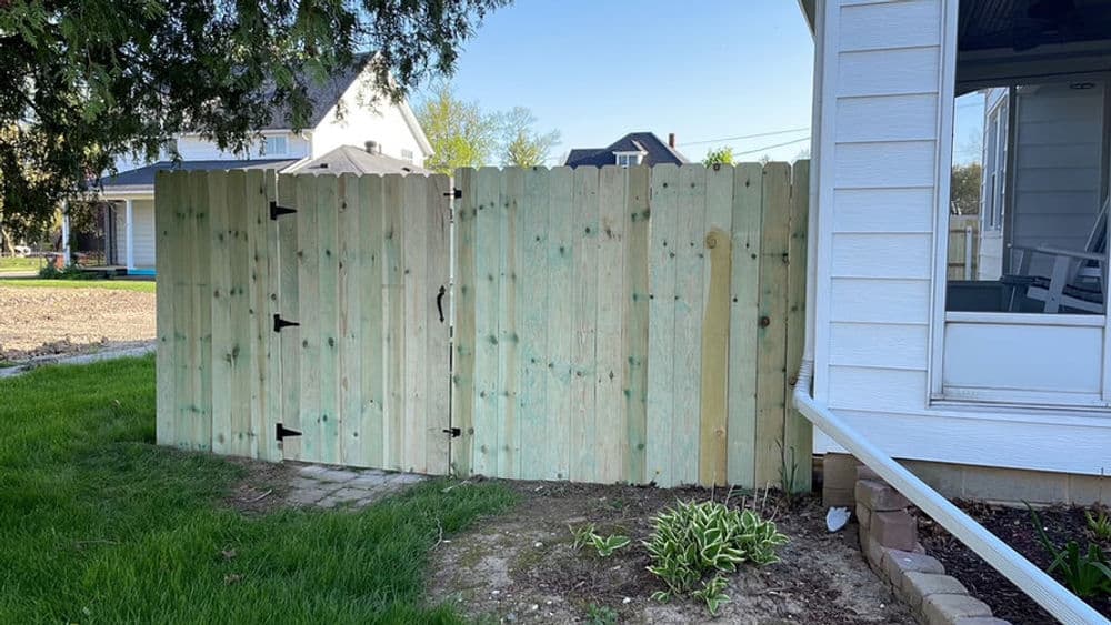 Wooden fence gate with black hinges beside a house and green lawn.