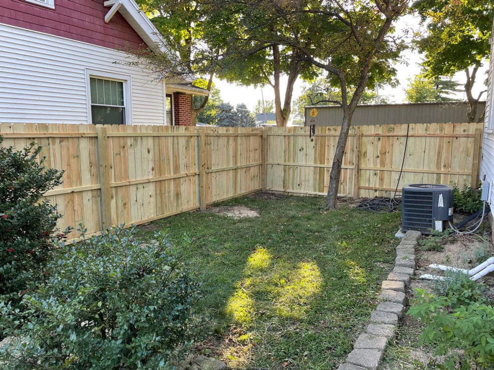 Wooden fenced yard with green grass, surrounded by trees and shrubs, sunny day.