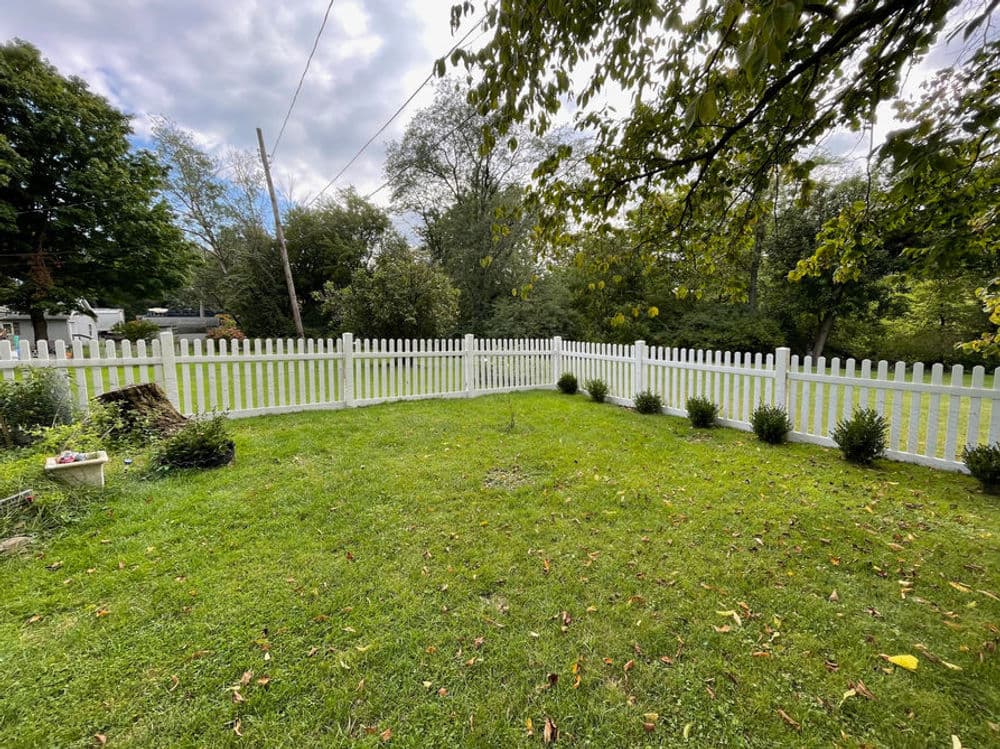 White picket fence enclosing a green yard with bushes and trees under a cloudy sky.