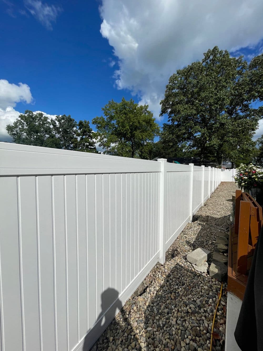 White vinyl fence along a gravel path under a blue sky with trees in the background.