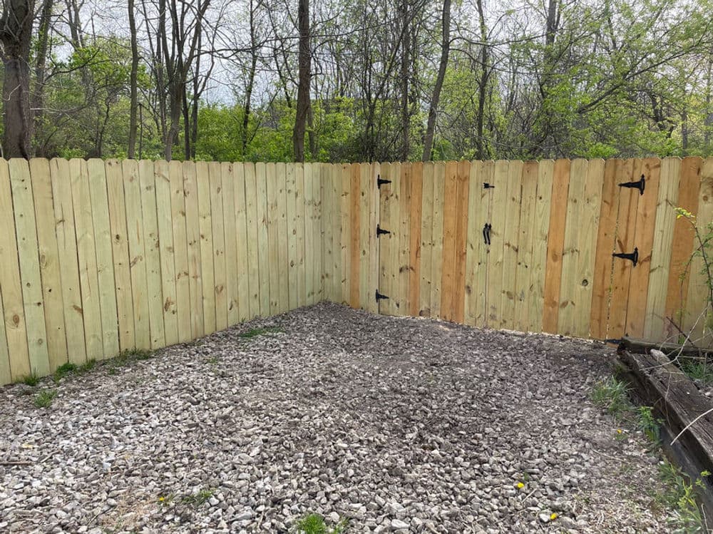 New wooden fence installation in a gravel area surrounded by trees.