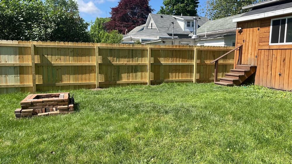 Newly built wooden fence surrounding a grassy backyard with a brick fire pit and wooden steps.