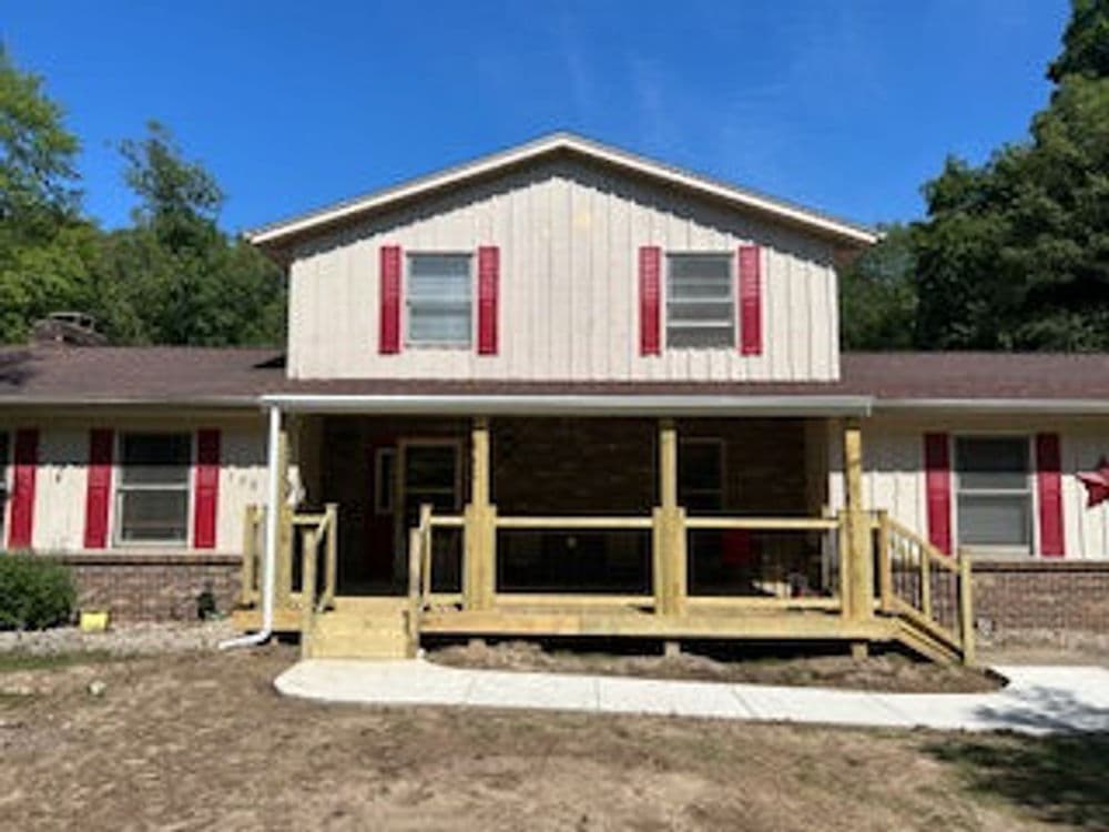 Two-story house with a front porch, red shutters, and a landscaped yard under clear blue sky.