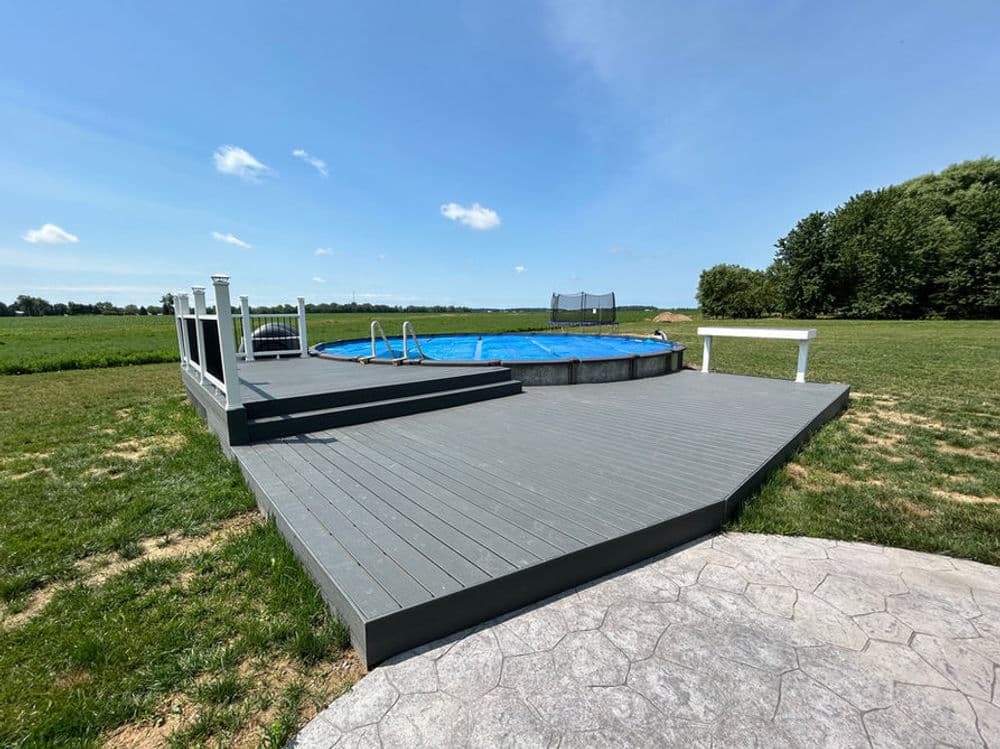 Above-ground pool on a wooden deck surrounded by green grass and trees under a clear blue sky.