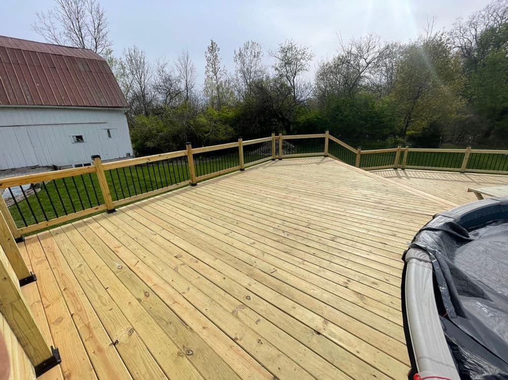Renovated wooden deck with railing and a hot tub, surrounded by greenery and trees.