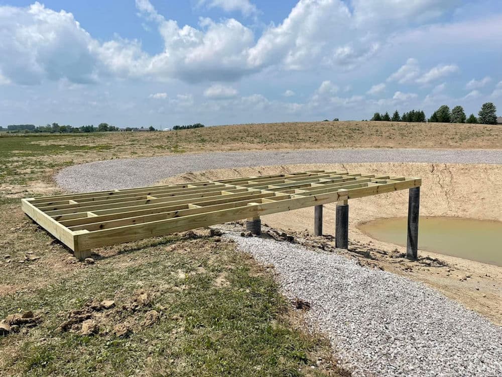 Wooden platform structure on stilts near a pond under a cloudy sky in an open landscape.