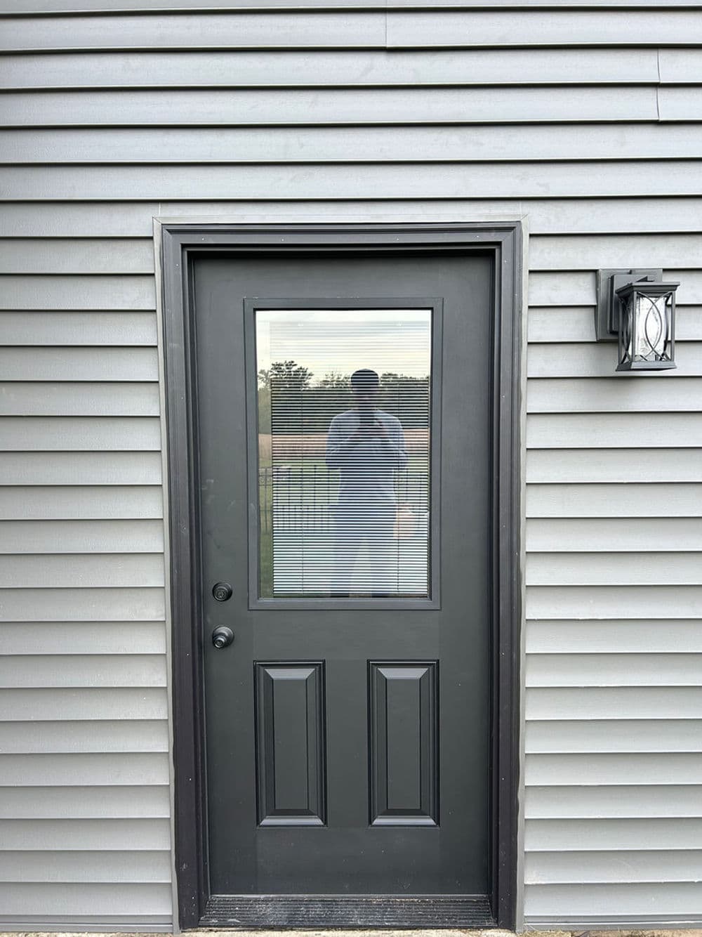 Exterior view of a modern black door with vertical blinds, set against a horizontal gray siding backdrop.