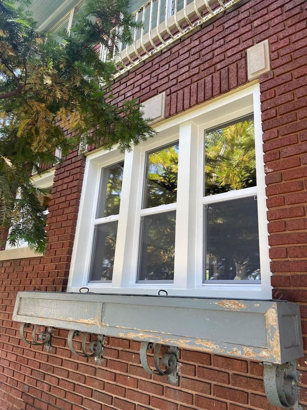 Brick wall with white window trim and a gray planter box beneath the window. Green tree reflections.