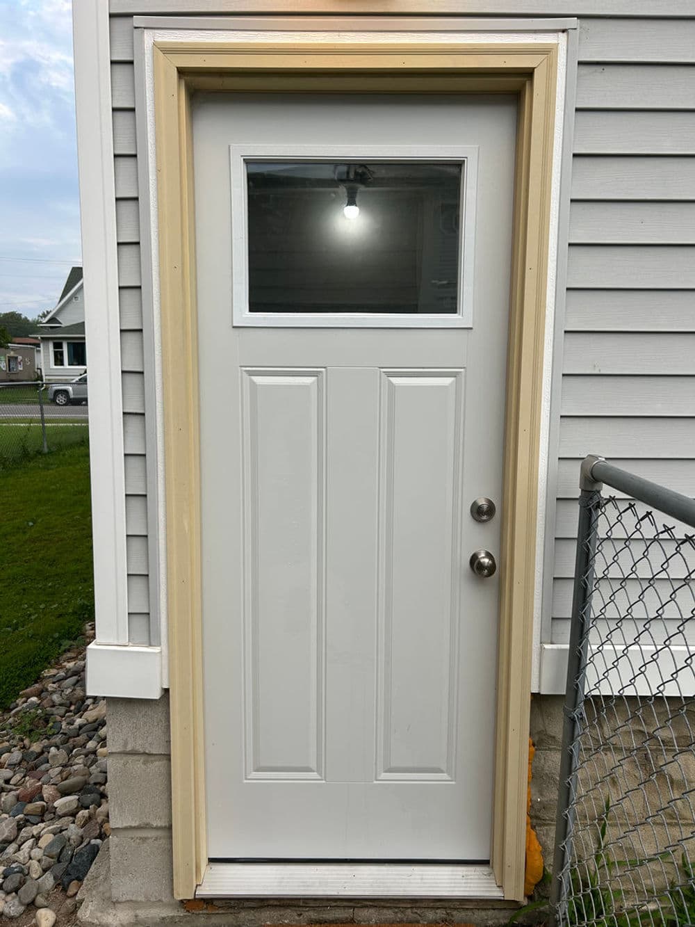 White exterior door with a small window, framed in beige, set against a gray siding background.